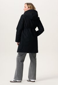 Vrouw die zijwaarts kijkt draagt een zwart hooded jasje, grijze wijde broek en witte sneakers met zwarte strepen tegen een effen achtergrond.