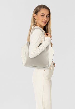 Sac fourre-tout en cuir gris au design spacieux, avec une texture douce et des coutures contrastantes, tenu par une personne portant un haut en rib blanc.