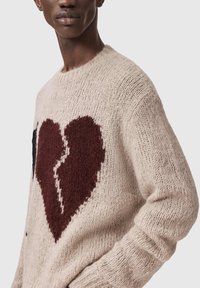 Πλεκτό πουλόβερ σε μπεζ χρώμα με μεγάλο σπασμένη καρδιά σε μπορντό και μαύρο. Διαθέτει στρογγυλή λαιμόκοψη και υφή νήματος.