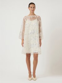 Γυναίκα που στέκει μέσα σε ένα άσπρο κομψό φόρεμα από λευκή δαντέλα με παγιέτες, διαθέτοντας μακριά διάφανα μανίκια και συνδυασμένο με κρεμ ψηλοτάκουνα παπούτσια.