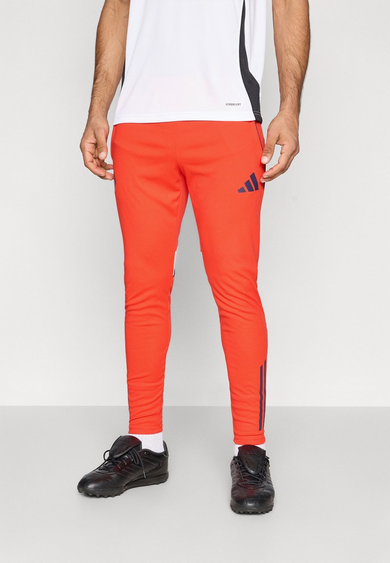 Pantalones deportivos rojos con diseño ajustado, con franjas azul marino y el logo de Adidas. Combinados con zapatillas deportivas negras y calcetines blancos.
