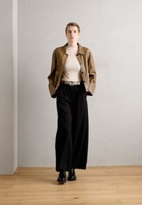 Braune Lederjacke über einem beigen gerippten Oberteil, kombiniert mit weiten schwarzen Hosen und schwarzen Stiefeln. Einfaches Design mit einem schlanken Gürtelakzent.