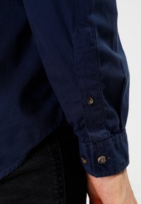 Tummansininen paita, jossa on napitettu hihansuu, jonka erityispiirteitä ovat sileä pinta ja jämäkkä muotoilu. Näyttää ompeluyksityiskohtia ja tummia nappeja.