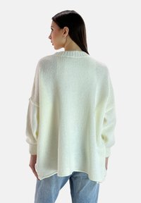 Weißer, übergroßer Strickpullover mit lockerer Passform, geripptem Halsausschnitt und seitlichen Schlitzen, kombiniert mit hellblauen Jeans. Weicher, strukturierter Stoff.