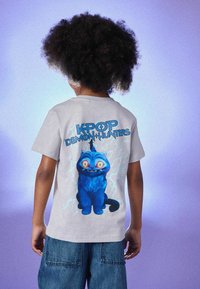 Graues T-Shirt mit einer blauen Cartoon-Katze, die große gelbe Augen und ein schelmisches Grinsen hat. Aufschrift: "KPOP DEMON HUNTERS."