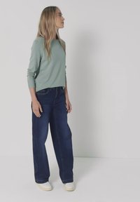 Frau mit langen blonden Haaren, die ein hellgrünes langärmliges Oberteil, dunkle weite Jeans und weiße Schuhe trägt, steht vor einem schlichten weißen Hintergrund.