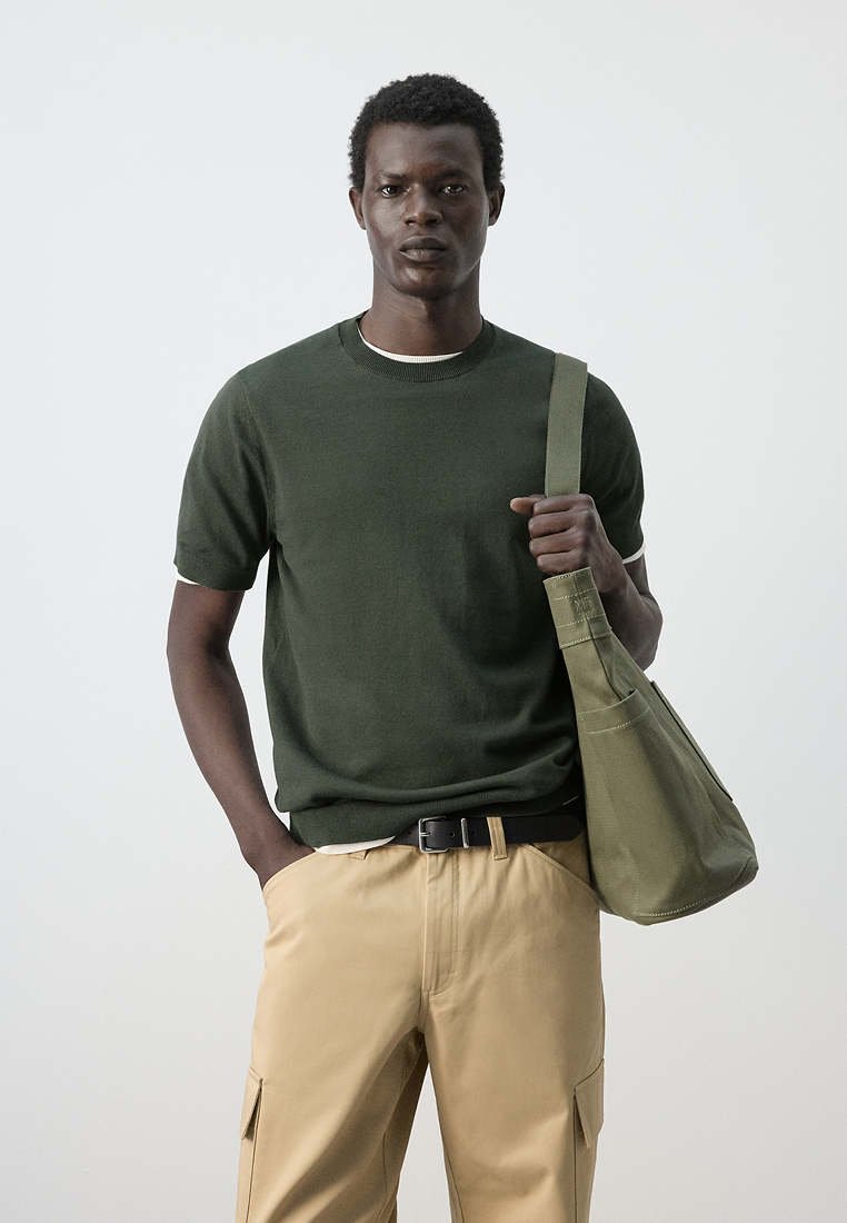 Homme portant une chemise à manches courtes vert foncé et un pantalon cargo beige, debout tenant un sac à bandoulière vert olive avec une main dans sa poche.