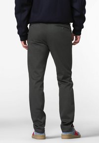 Persoon in een donkergroene broek, zwart sweater en blauwe sneakers met rode accenten, die met de rug naar de camera staat op een effen witte achtergrond.