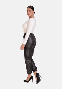 Aansluitend wit longsleeve-shirt met geplooide voorkant, gedragen met zwarte leren joggingsbroeken met elastische boorden en zijzakken. Zwarte hakken.
