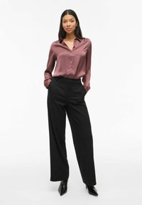 Blusa bordeaux in satin con colletto e bottoni frontali abbinata a pantaloni neri a gamba larga a vita alta. Modella in tacchi a punta neri.