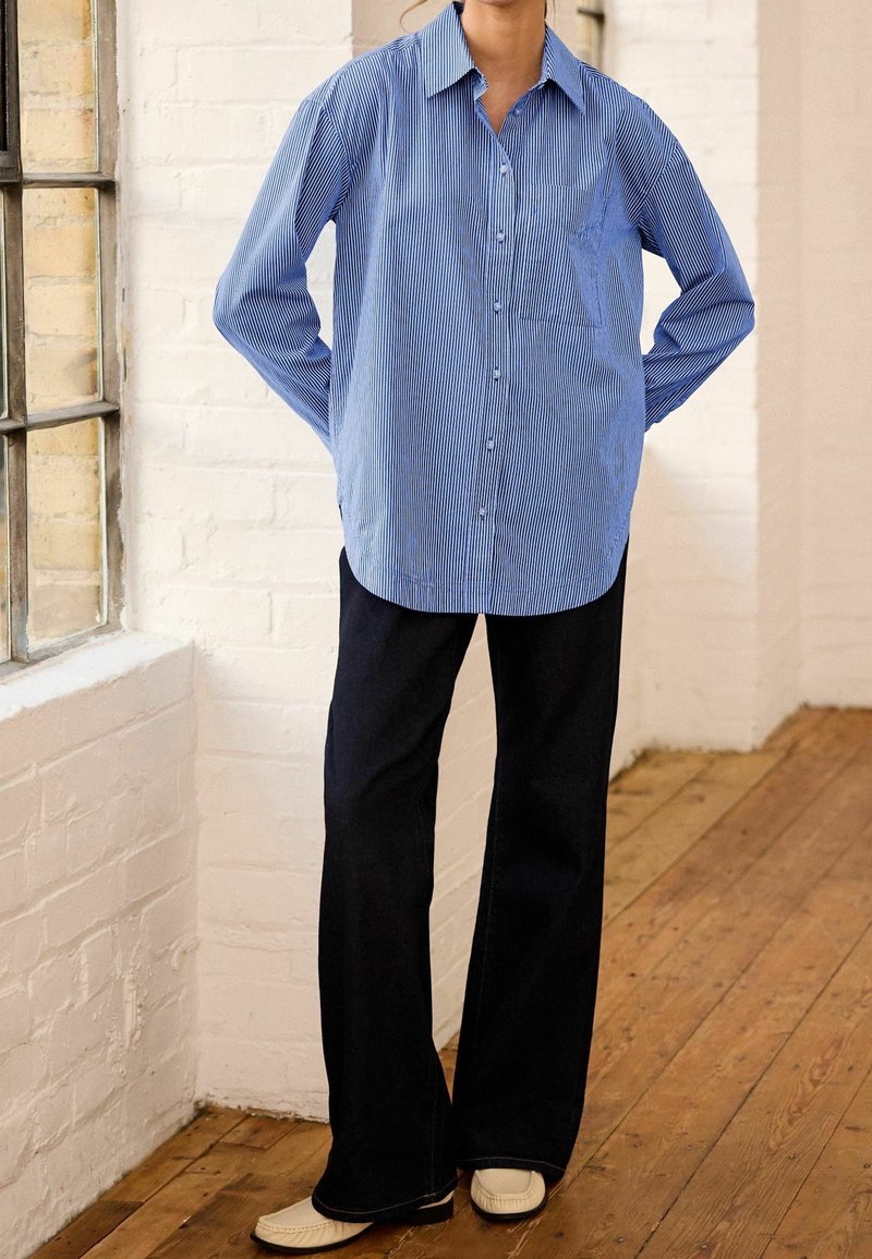Personne portant une chemise rayée bleu et blanc à boutons, un pantalon noir à jambes larges et des chaussures beige clair, debout sur un sol en bois près d'une fenêtre.