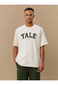 Cremefarbene Baumwoll-T-Shirt mit fetter schwarzer "YALE" Textgestaltung, lockere Passform und kurzen Ärmeln. Kombiniert mit dunkelgrünen Jogginghosen.