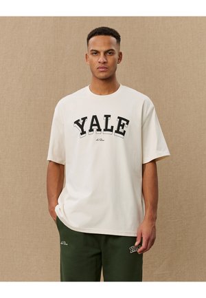 Tricou din bumbac de culoare crem, cu imprimare îndrăzneață în negru a textului "YALE", croială lejeră și mâneci scurte. Asortat cu pantaloni de trening verzi închis.