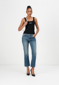 Schwarzes, tailliertes Tanktop mit einem Logo, blaue Jeans mit einem gekürzten, fransigen Saum und schwarze, spitze Absätze, ergänzt durch eine kleine weiße Handtasche.