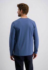 Mann von hinten zu sehen, der ein blaues Langarmhemd und dunkle Hosen trägt, vor einem schlichten hellen Hintergrund stehend.