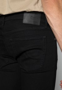 Zadní pohled na osobu v černých džínách Jack & Jones, s béžovou košilí částečně viditelnou nad pasem.