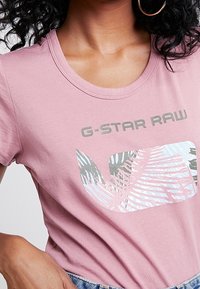 Růžové bavlněné tričko s grafickým designem obsahujícím palmové listy a text "G-STAR RAW" v šedé barvě. Kulatý výstřih a jemná textura.