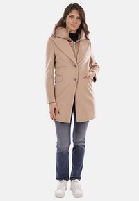 Cappotto beige aderente con cappuccio ampio, chiusura con bottoni e due tasche frontali. Abbinato a jeans blu e scarpe da ginnastica bianche. Tessuto liscio.