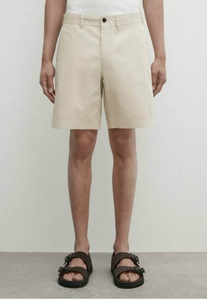 Homme portant un short beige jusqu'aux genoux et des sandales marron à double lanière, debout devant un fond clair uni.