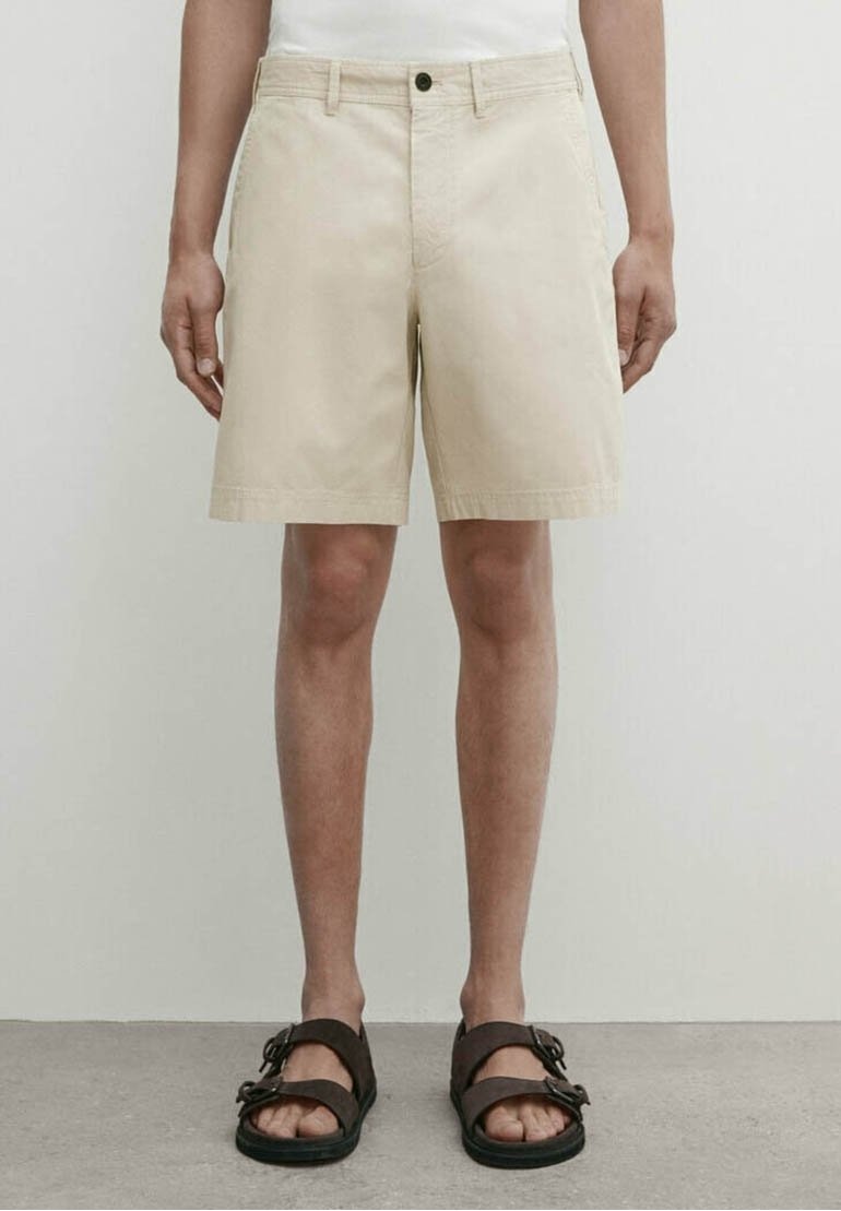 Homme portant un short beige jusqu'aux genoux et des sandales marron à double lanière, debout devant un fond clair uni.
