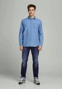 Camisa chambray azul claro com colarinho abotoável e bolso no peito, usada com jeans azul escuro e ténis cinzentos com detalhes brancos.