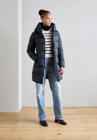 Chaqueta de plumas azul marino con capucha, con textura acolchada, llevada sobre un suéter a rayas y combinada con jeans de color azul claro y botas negras de tobillo.