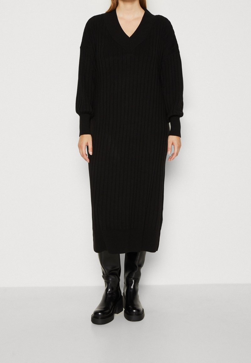 Personne portant une robe longue noire en maille côtelée avec manches longues et col en V, associée à des bottes noires brillantes montantes jusqu'aux genoux.