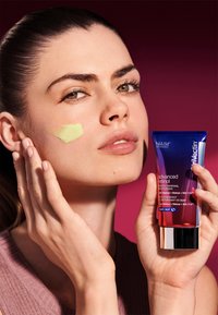 Il tubo del moisturizer avanzato a base di retinolo NIA114, con un design blu e viola, è tenuto accanto a un viso con una crema di colore verde chiaro applicata. Texture liscia.