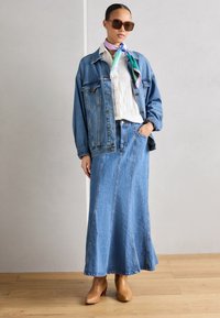 Denim jack over een wit shirt, gecombineerd met een lange denim rok. Bruine haklaarzen tot de enkel, een kleurrijke zijden sjaal om de hals geknoopt.