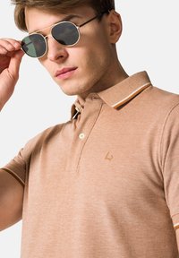 Brązowa koszulka polo z fakturowanym splotem, kontrastowym kołnierzykiem w białe i brązowe paski, z małym logo na piersi. Srebrne okulary przeciwsłoneczne.