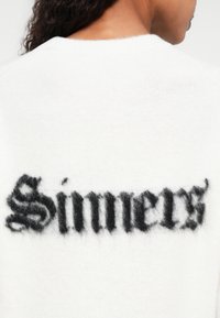 Biały sweter z puszystym czarnym nadrukiem na plecach z napisem "Sinners". Miękka tekstura i klasyczny krój z okrągłym dekoltem.