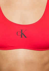 Rød sports-bh laget av glatt stoff, med rund halslinning, enkelt design og en kontrastfarge sort "cK"-logo på fremsiden.