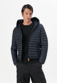 Navy puffer jacket with horizontal quilted pattern, hood, and a front zipper featuring an orange pull tab. Worn over a black shirt.