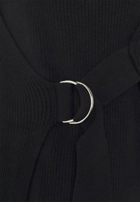 Tejido negro acanalado con un acento de anillo plateado. Presenta un diseño envolvente con un acabado suave y texturizado, y un detalle de correa ajustable.