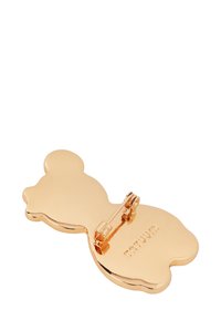 Auksinė segė meškos formos, blizgiu paviršiumi ir su smeigtuko uždarymu gale. Ant nugarėlės išgraviruotas logotipas, kraštai lygūs ir apvalūs.