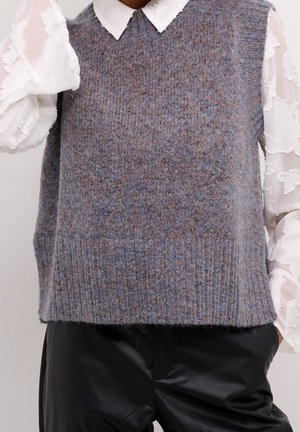 Harmaa neulottu hihaton liivi valkoisen, teksturoidun, kauluksellisen pitkähihainen paidan päällä, yhdistetty mustiin nahkahousuihin.