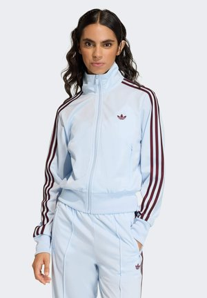 Kobieta w jasnoniebieskiej kurtce i spodniach dresowych Adidas z bordo paskami i logo, stojąca z jedną ręką w kieszeni.