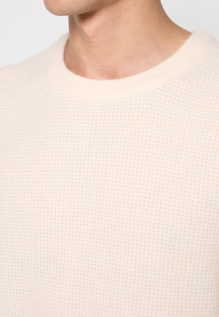 Hell gefärbter, strukturierter Strickpullover mit rundem Halsausschnitt. Eine Nahaufnahme hebt das Gewebe und das weiche Erscheinungsbild des Stoffes hervor.