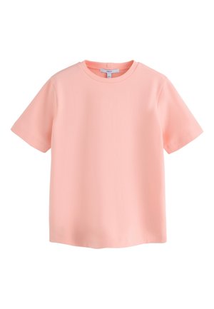 Kvinde med langt mørkt hår iført en pink skjorte og denimshorts står på en træbro ved havet, armene krydsede, kigger mod venstre.