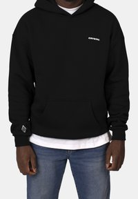 Mustat hupparit, jotka on valmistettu pehmeästä kankaasta, sisältävät etutaskun, resorikääntömainokset ja valkoista logo-tekstiä rinnassa. Kerrospukeutuneena valkoisen paidan päälle.