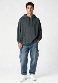 Felpa grigia scura con tasca a marsupio e cappuccio con stringa, abbinata a jeans cargo blu chiaro e sneaker bianche. Tessuto morbido e casual.