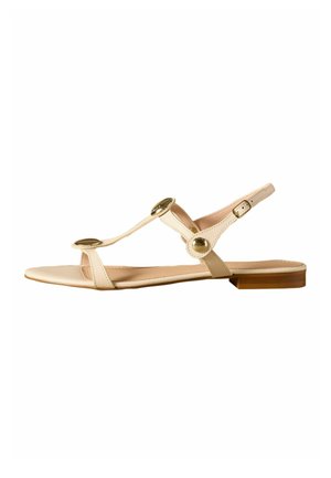 Beige flad sandal med tynde stropper, guld cirkulære pynt, en lille spænde og en lav træhælc.