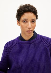 Dunkelvioletter Strickpullover mit gerippter Textur und Rundhalsausschnitt, über einem dunkelblauen Hemd mit Kragen getragen. Verfügt über Raglanärmel.