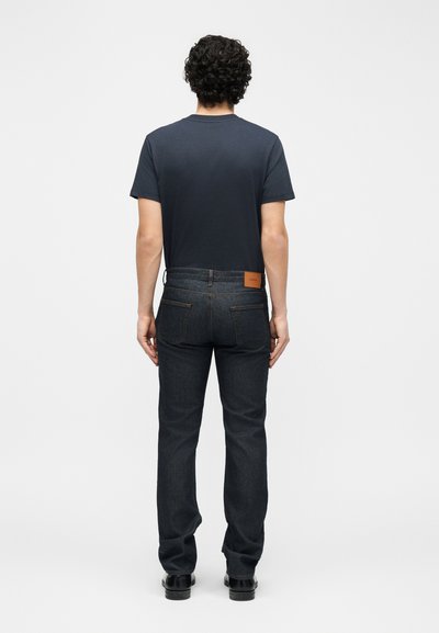 Personne aux cheveux bouclés portant un t-shirt sombre, un jean en denim foncé et des chaussures noires, debout dos à la caméra sur un fond blanc uni.