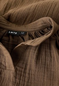 Smeđa lanena bluza s V-izrezom i skupljenim detaljima, etiketa s natpisom "LMTD" i veličinom "XXS." Tkanina ima suptilan teksturirani uzorak.