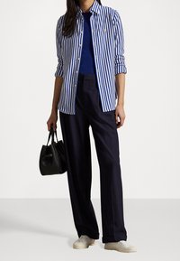 Niebieska w paski koszula z guzikami noszona na niebieskiej bluzce, zestawiona z luźnymi granatowymi spodniami i białymi trampkami, trzymająca czarną torebkę.