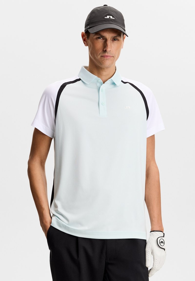 Homme portant une casquette noire, un polo bleu clair et blanc avec des accents noirs, un pantalon noir, et un gant de golf blanc à la main droite, debout avec une main dans la poche.