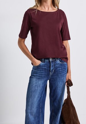 Frau trägt ein burgunderfarbenes Kurzarmoberteil und blaue Jeans, hält eine braune Wildlederhandtasche in einer Hand und hat die andere Hand in der Tasche.