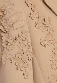 Țesătură bej cu detalii florale aplicate texturate. Florile sunt tridimensionale, cu accente cusute, creând un design stratificat.
