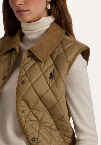 Vadderad brun väst med kordflätselkrage, med tryckknappar och en liten broderad logotyp. Bärs över en ljusbeige polotröja.